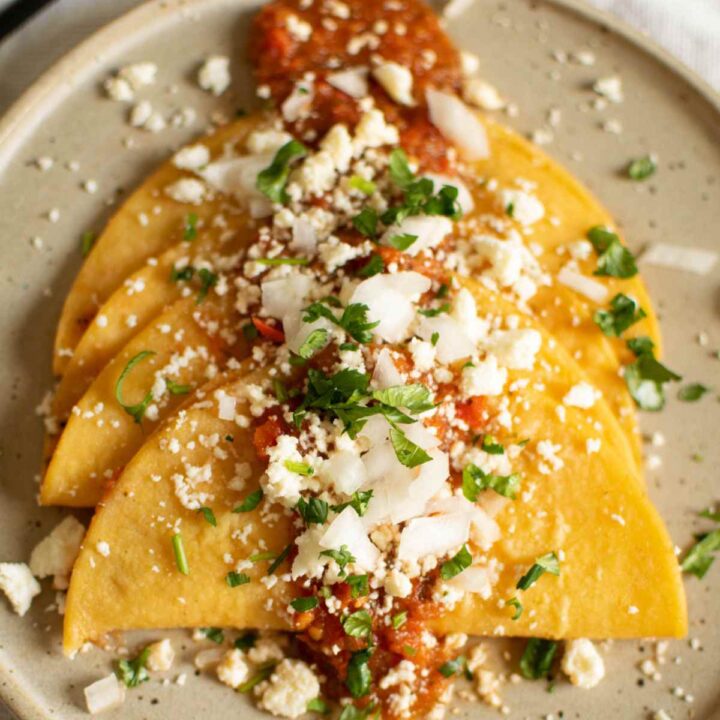 3 entomatadas on a plate bathed in tomato sauce with queso fresco crumbled over the tops of them.