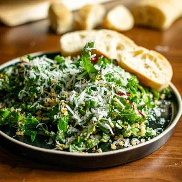 featured image for swiss chard salad; it shows swiss chard salad tossed in parmesan and breadcrumbs served on a plate with three slices of baguette
