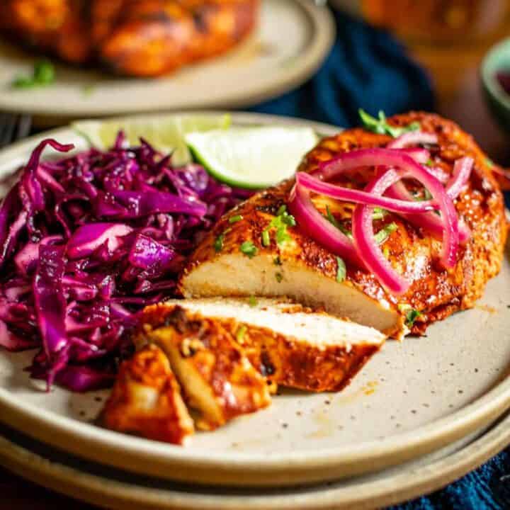 A plate of half sliced achiote chicken, topped with sliced pickled red onions and purple coleslaw.