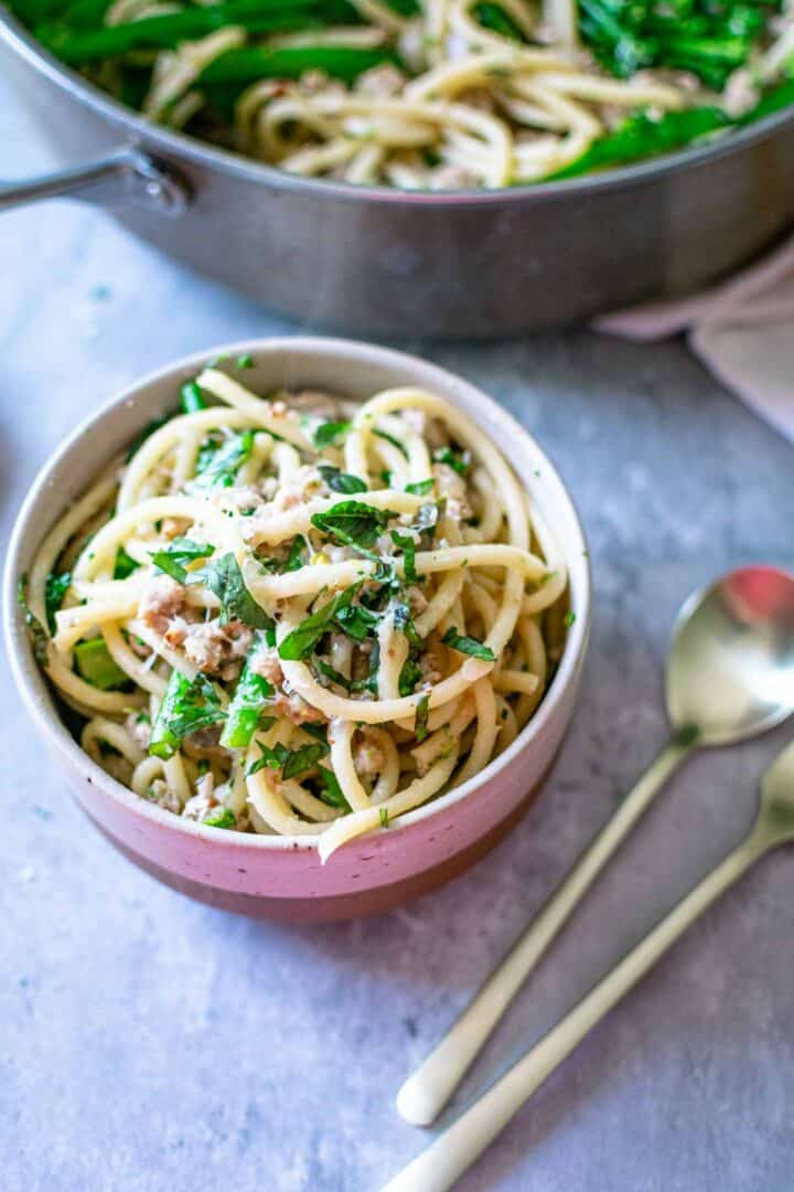A bowl of sausage pasta with silverware and a pan of pasta in the background.