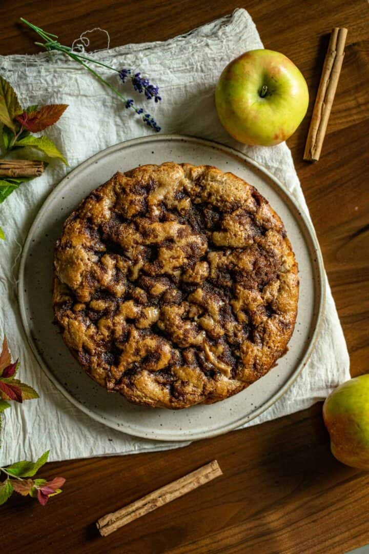 an overhead shot of french apple yogurt cake dimpled with cinnamon sugar crunch topping and surrounded by sticks of cinnamon, an apple, lavender, and some red green leaves from a bush on a walnut table.