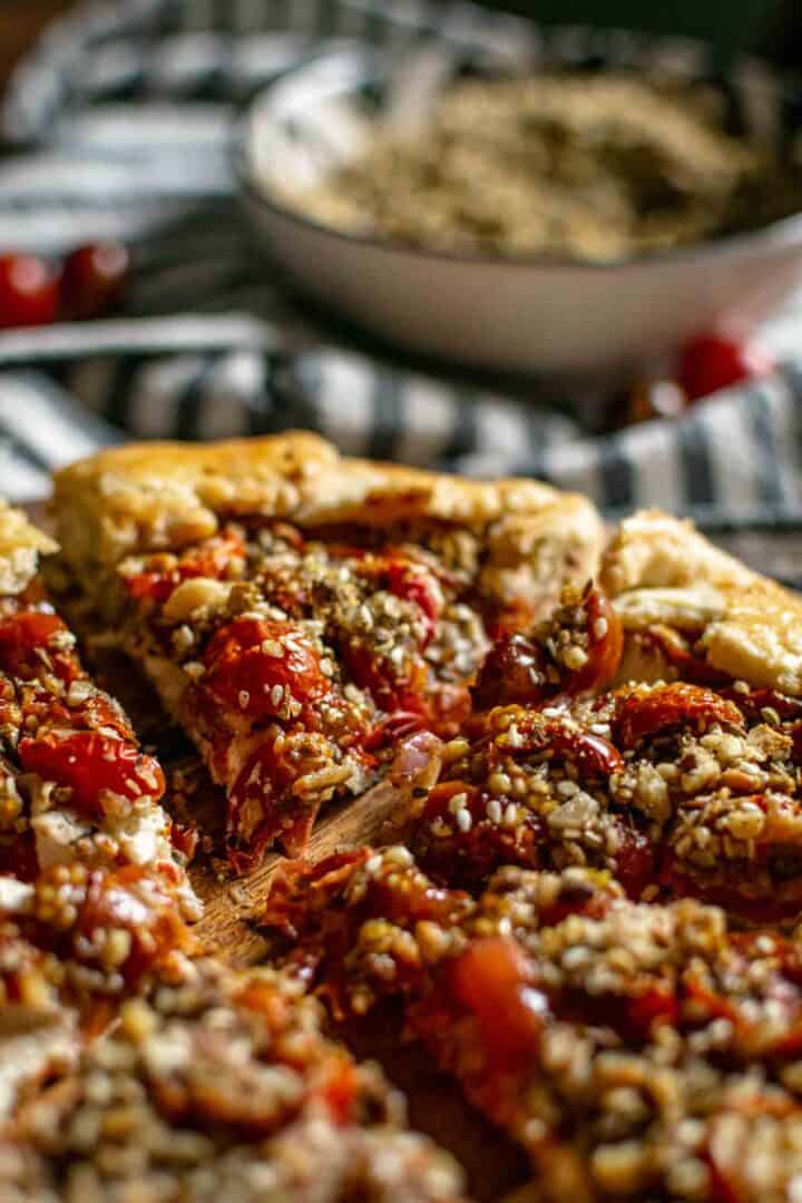 Slices of cherry tomato galette on a wooden serving tray with a backgroundt hat includes a striped tea towel and a bowl of dukkah.