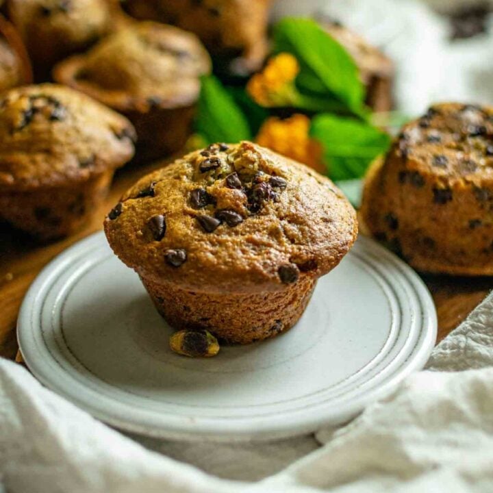 Featured image of pistachio chocolate chip muffin, which is on a cream colored tea saucer and has pictures of muffins in the back along with two small orange flowers, blurred out with bokeh.