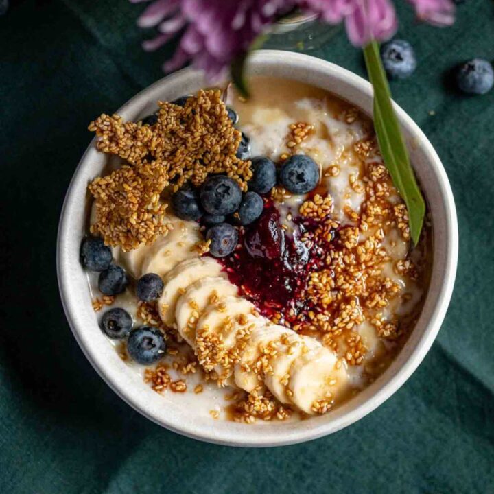 Overhead shot featuring blueberry tahini oatmeal porridge with sesame brittle, sliced banana, blueberry compote in a white bowl on a green background, under a few blurred purple flowers.