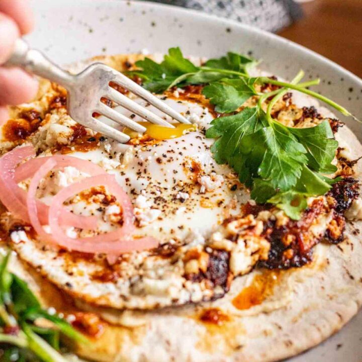 fork piercing feta fried egg, which is sitting on top of hummus and a pita and garnished with a tangle of pickled red onions and a few sprigs of parsley.