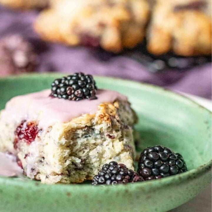 blackberry scone on a green plate surrounded by two other whole blackberries with a cooling tray of scones sitting on purple fabric in the blurred background.