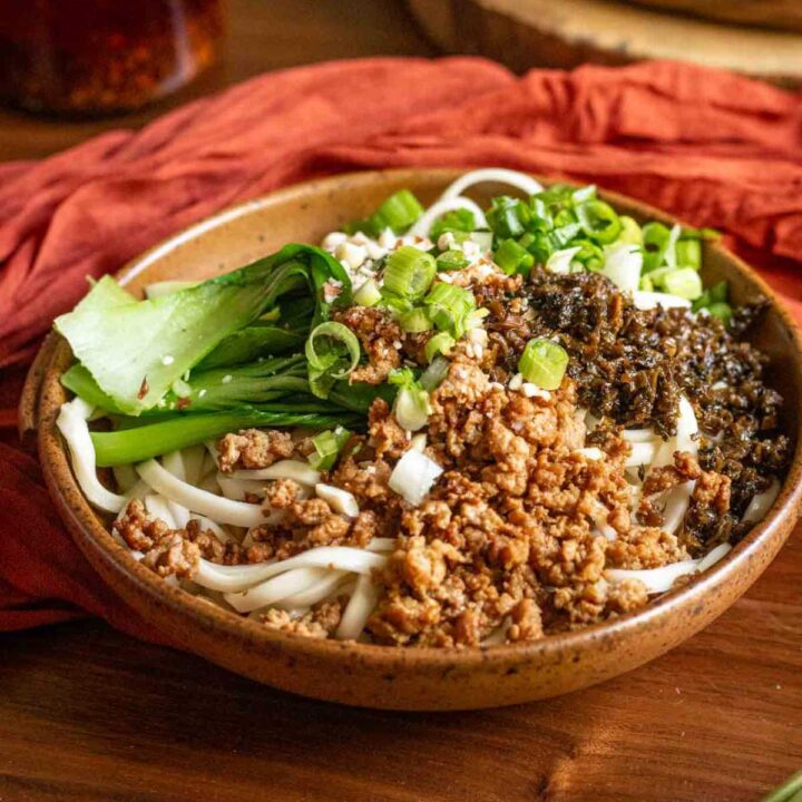 A bowl of yibin ran mian, or burning noodles, pictured before its been tossed in sauce and featuring toppings of minced pork, mustard pickle, green onions peanuts and shanghai bok choy.