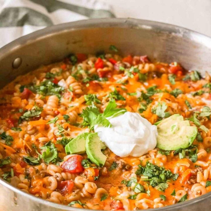 A pot of cheesy taco pasta with avocado slices, sour cream, and cilantro garnishes.