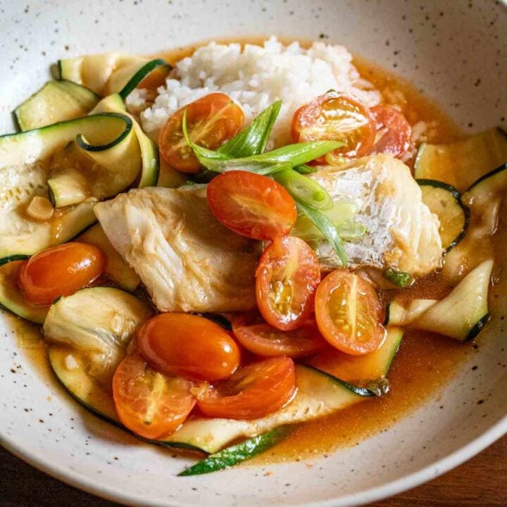 Overhead shot of cod sitting on top of a bed or rice, topped with a few cherry tomato halves and scallions with zucchini ribbons and a red broth.