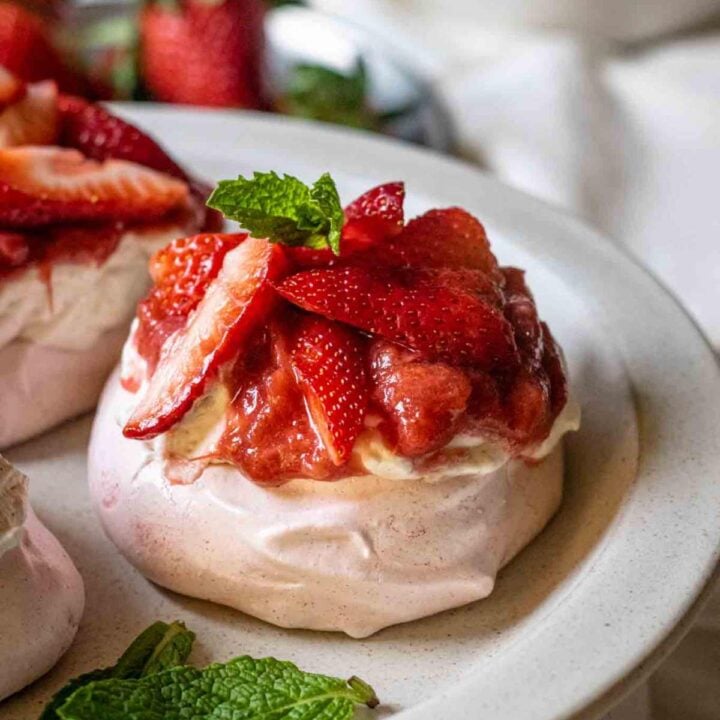 A close up front angled shot of a pink pavlova nest topped with whipped cream, a red sauce and sliced strawberries surrounded by a few leaves of fresh mint and a plate piled high with strawberreies against a white linen background.