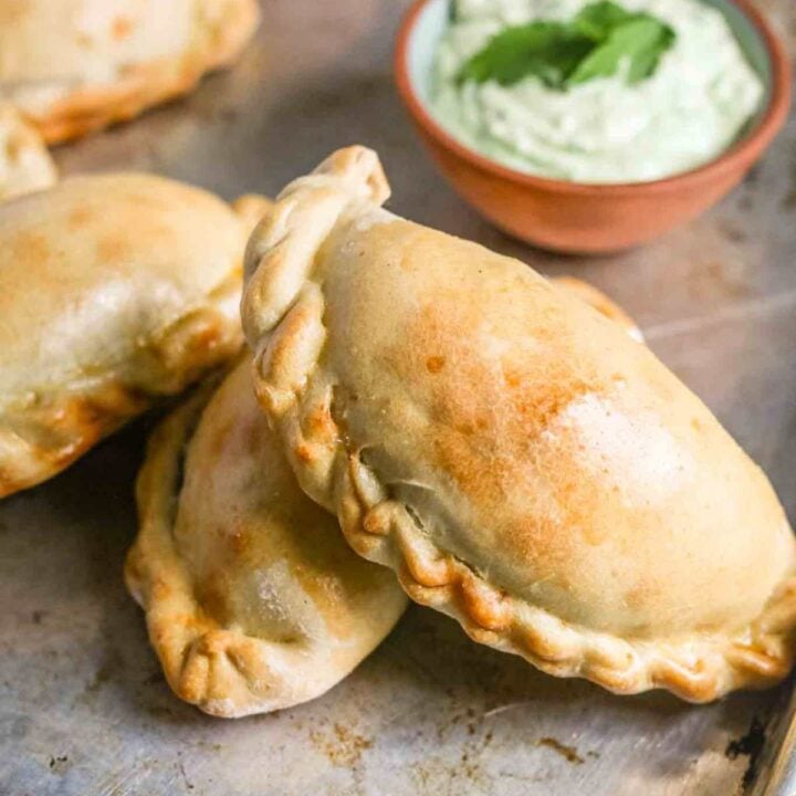 Empanadas on a cookie sheet, arranged sort of stacked and splayed out for the reader with a pinch bowl of avocado crema in the background.