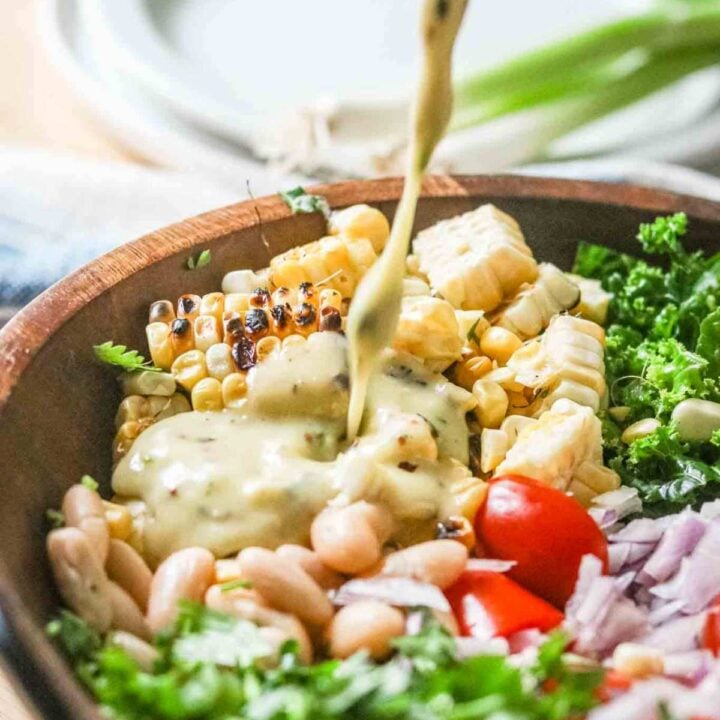 Dressing being poured over a bowl of salad that includes charred corn, cherry tomatoes, kale, white beans, cilantro and red onions.