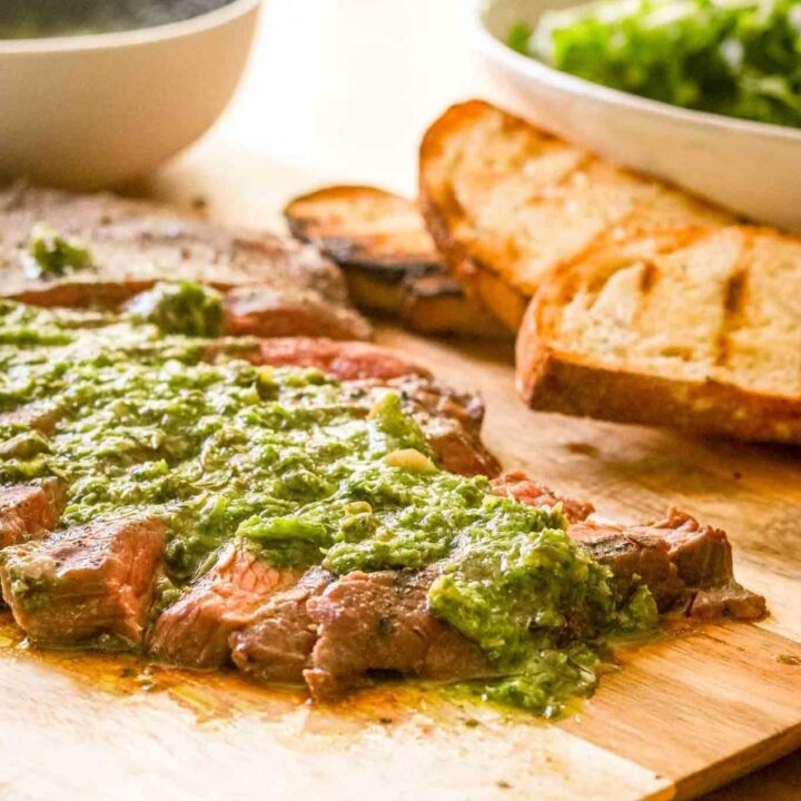 Sliced flank steak cooked in the sous vide on a wood cutting board with a green sauce and grilled bread and salad.