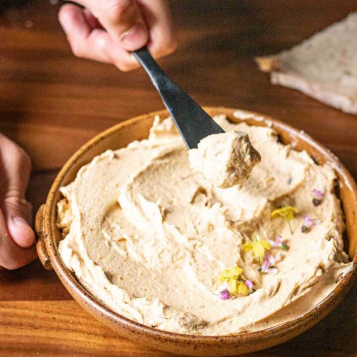 One hand scooping up bottarga pate on a black butter knife and another hand holding the edge of a brown clay serving dish.