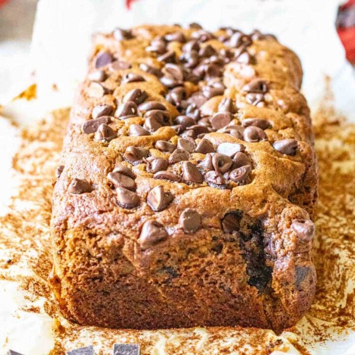 The whole loaf of chocolate chunk banana bread on the parchment paper is was cooked in with some chocolate chunks in the foreground and red cloth in the background.